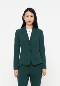 Tmavě zelený upravený blazer s jedním knoflíkem, s fazonkovými klopami a bočními kapsami. Nosený přes bílou košili s logem.