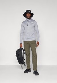 Grå langermet zip-up topp, olivengrønne cargo bukser, svart bøttehatt, svart ryggsekk og mørke sko, med merkevarelogo på toppen og buksene.