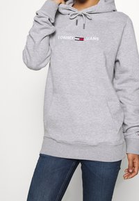 Felpa grigia con cappuccio, dotata di tasca frontale e cordoncino. Logo ricamato "TOMMY JEANS" in bianco, con un accento a strisce colorate.