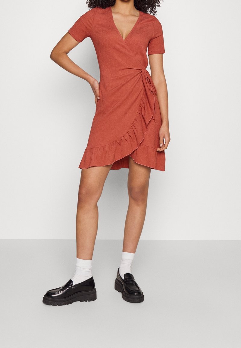 Femme portant une robe portefeuille rouille à manches courtes avec un volant à l’ourlet, des chaussettes blanches et des mocassins noirs épais, debout sur un fond clair uni.