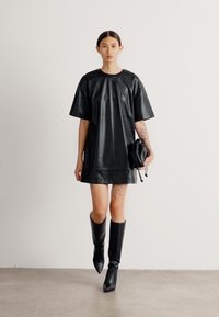 Vestido de couro preto com mangas curtas e decote redondo, combinado com botas pretas até ao joelho. O modelo segura uma pequena bolsa preta.