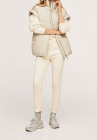 Chaleco acolchado beige con cuello alto, llevado sobre un suéter crema y pantalones deportivos de color claro. Combinado con calcetines grises y zapatillas claras.