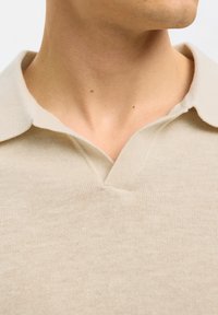 Polo tricot beige avec col en V et tissu texturé. Le design met en avant un style épuré et minimaliste.