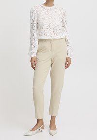 Top in pizzo a maniche lunghe bianco con motivo floreale, abbinato a pantaloni beige su misura. Il modello indossa décolleté bianchi a punta e con cinturino sul tallone.