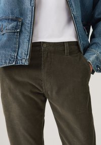 Corduroy trousers in dark olive green with a soft texture, featuring a flat front and side pockets, paired with a light denim jacket.