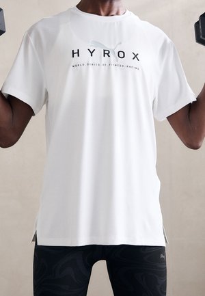 Persona levantando mancuernas, vistiendo una camiseta blanca de Hyrox fitness y mallas negras con estampado sobre fondo neutro.