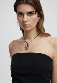 Silberner Anhänger mit strukturiertem Design an einem schwarzen Band, getragen von einer Person in einem schwarzen trägerlosen Top. Glatte, reflektierende Oberflächen.