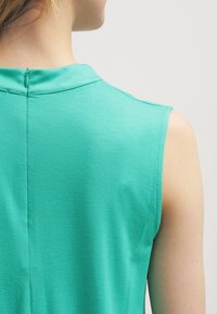 Haut sans manches vert en tissu lisse. Présente un col montant et un détail de fermeture éclair au dos. La texture semble légère et respirante.