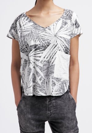 Mujer que lleva una blusa de cuello en V de manga corta con estampado tropical en blanco y negro, con un bolsillo delantero blanco y jeans de lavado oscuro.