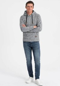 Sudadera gris melange con cordones, bolsillo canguro frontal y una pequeña etiqueta con el logo, combinada con jeans azules y zapatillas blancas.