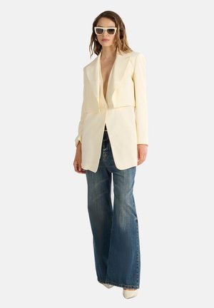 Cremfarbener Blazer mit tiefem V-Ausschnitt und strukturierten Schultern, kombiniert mit lässigen blauen Jeans mit Schlag und weißen High Heels.