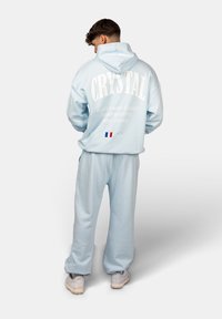 Himmelblauer Hoodie mit weißem Text auf der Rückseite, der "CRYSTAL" und Adressdetails von Paris zeigt. Kombiniert mit passenden himmelblauen Hosen.