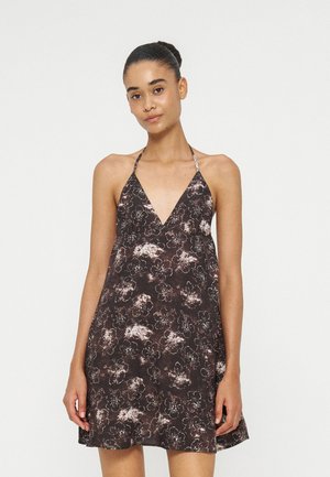 Femme avec les cheveux en chignon portant une robe marron fleurie sans manches avec un profond décolleté en V sur un fond clair uni.
