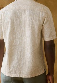 Camisa de manga corta en color beige claro, confeccionada en una tela texturizada que presenta un intrincado patrón de hojas. Bajo redondeado con costuras visibles.