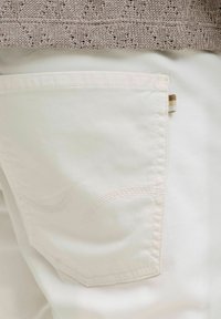 I pantaloni bianchi presentano una tasca posteriore con una parte superiore arrotondata, sottili dettagli di cucitura e una piccola etichetta in tessuto sul lato.