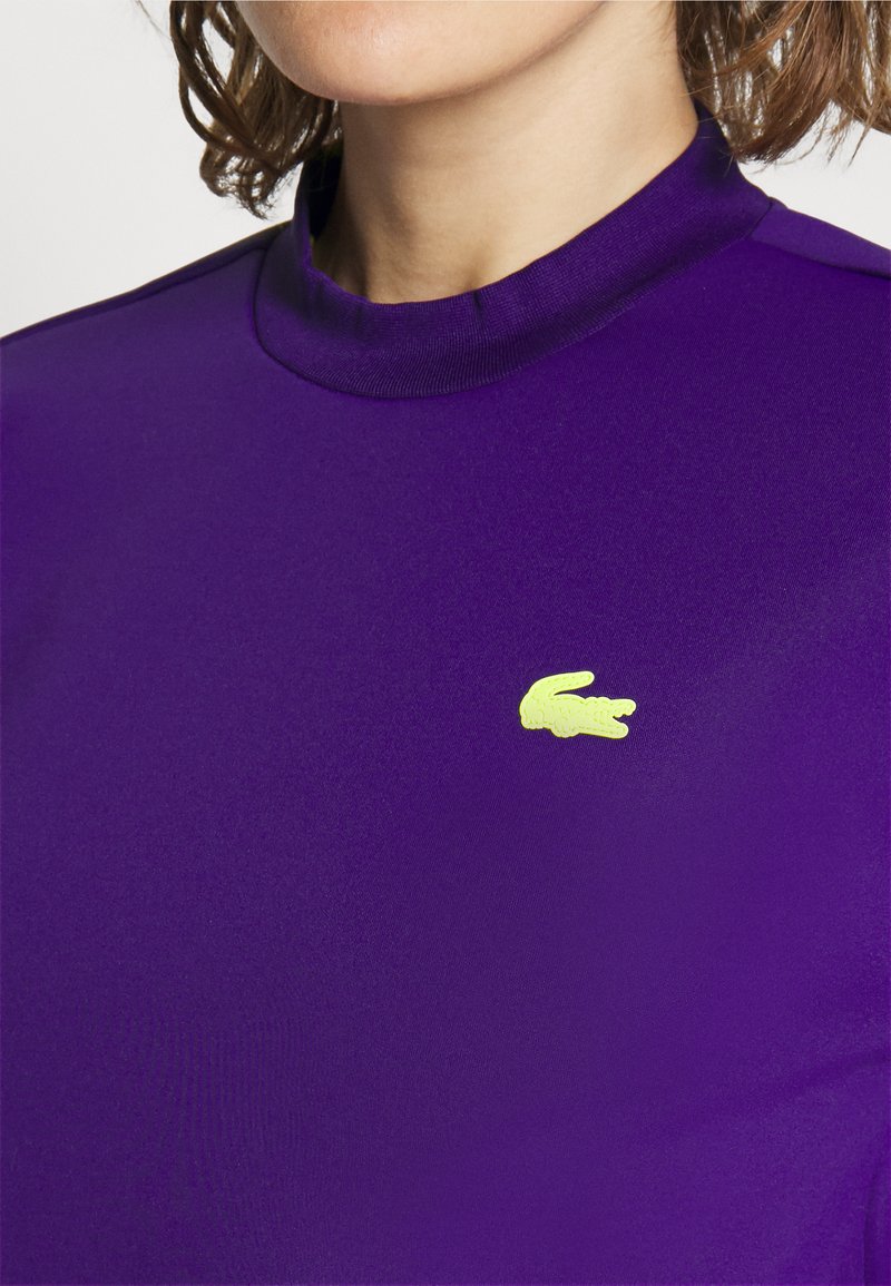 Haut à manches longues violet avec un col montant, en tissu lisse. Présente un petit logo de crocodile jaune sur la poitrine.