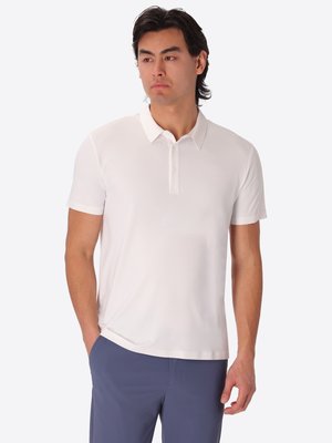 Uomo in piedi con le mani nelle tasche, indossa una polo bianca a maniche corte e pantaloni blu, guarda verso sinistra.