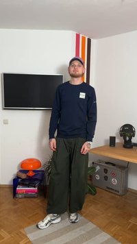 Maglietta a maniche lunghe blu navy con logo, pantaloni larghi verde oliva e scarpe da ginnastica argento. Sullo sfondo una lampada arancione e un tavolo di legno.