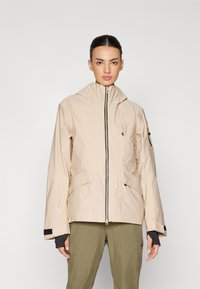 Beige vanntett jakke med høy krage, frontglidelås og to skrå lommer; har svarte detaljer og justerbare ermer.