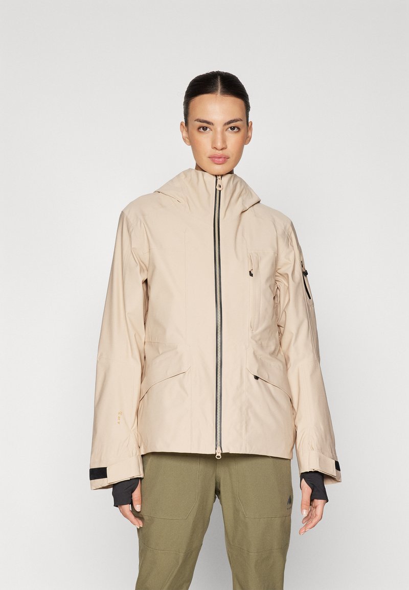 Beige vanntett jakke med høy krage, frontglidelås og to skrå lommer; har svarte detaljer og justerbare ermer.