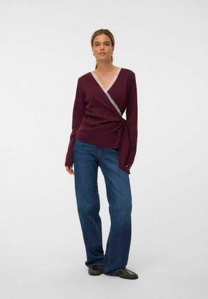 Frau trägt einen burgunderfarbenen Wickelpullover mit Spitzenbesatz, blaue Jeans mit weitem Bein und schwarze Schuhe, steht vor einem schlichten weißen Hintergrund.