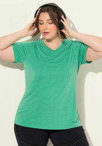 Zelené krátké tričko s výstřihem do V vyrobené z měkké látky, se uvolněným střihem a hladkou texturou, v kombinaci s černými sportovními kalhotami.
