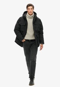 Sort pufferjakke med hætte og frontlommer, båret over en lys grå rullekravebluse og mørke jeans. Sorte støvler fuldender outfittet.