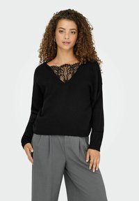 Zwarte v-hals trui met kanten detail bij de halslijn, lange mouwen en ribboordmanchetten, gecombineerd met grijze geplooide broek.