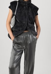 Black faux fur vest with high collar, paired with shiny grey satin pants. The outfit features a small silver handbag. Smooth textures, modern design.