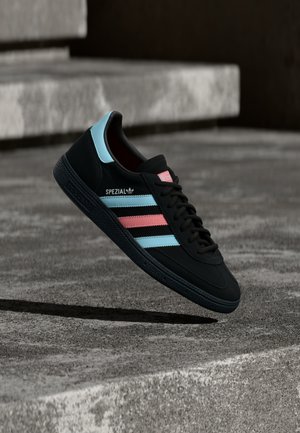 Μαύρο αθλητικό παπούτσι Adidas με μπλε και ροζ ρίγες, με το κείμενο «SPEZIAL» στο πλάι, αιωρούμενο πάνω από τσιμεντένια σκαλοπάτια σε υπαίθριο χώρο.