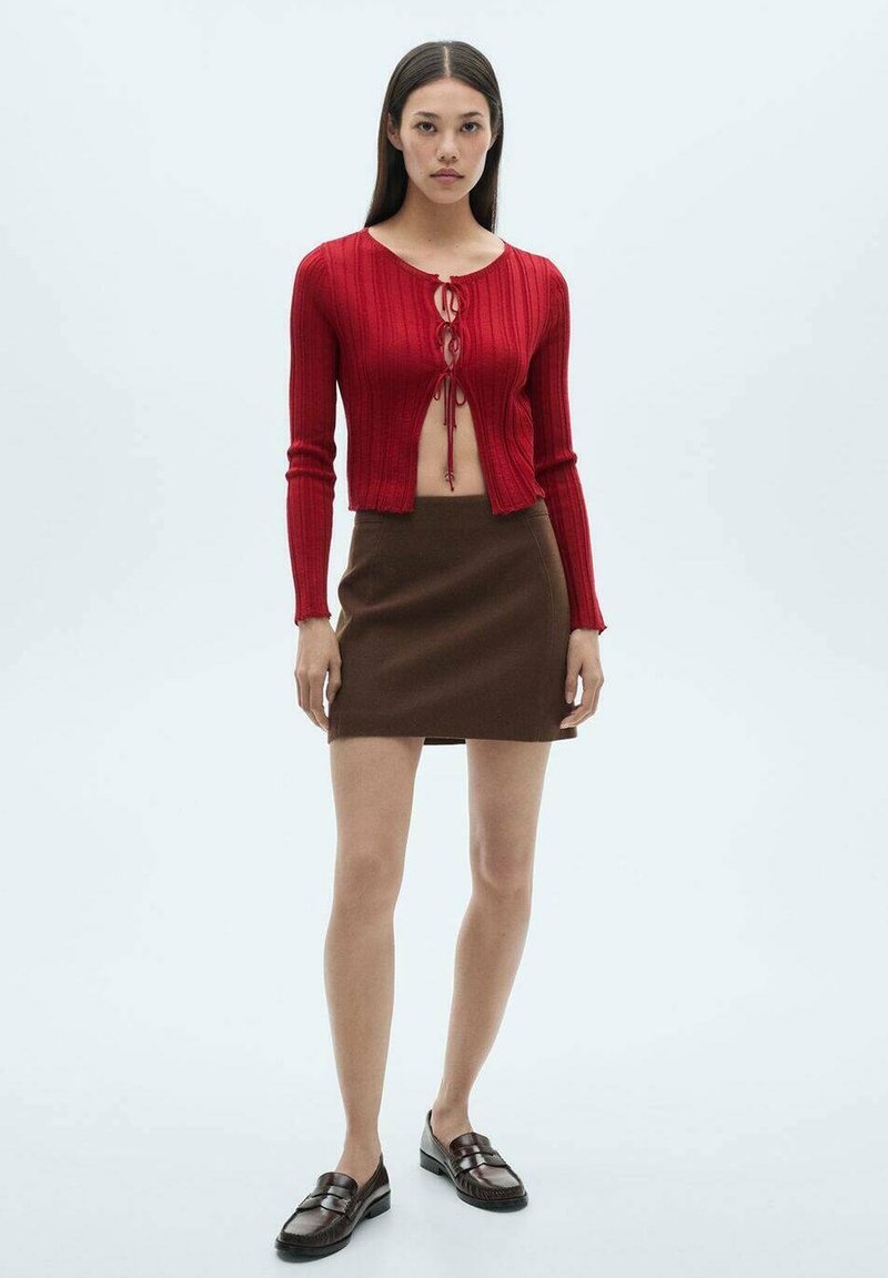 Rød ribbet cropped cardigan med snøre i front, parret med en brun mini nederdel. Modellen har mørkebrune slip-on sko på.