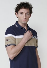 Sötétkék, bézs és fehér csíkos pólóing, gallérral, ujjon logóval és kontrasztos mandzsettával. Puha anyag, testhezálló fazon.