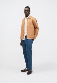 Hellbraune Baumwolljacke mit Knopfverschluss und Taschen, getragen über einem hellen karierten Hemd, kombiniert mit blauen Jeans und dunklen Lederschuhen.