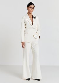 Blazer blanc avec un décolleté en V profond et une taille ceinturée, associé à un pantalon évasé. Présente un accent floral noir sur le blazer. Escarpins noirs.