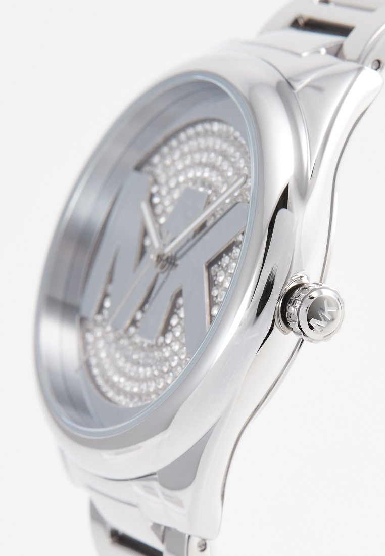 Zilveren roestvrijstalen horloge met een grote wijzerplaat versierd met kristallen, waarop de letters "MK" en een getextureerd logo zijn afgebeeld. Ronde vorm, gepolijste afwerking.