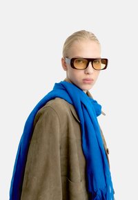 Przesadnie duże brązowe okulary przeciwsłoneczne z żółtymi soczewkami, szare zauszniki; niebieski teksturowany szalik opadający na beżowy zamszowy płaszcz. Gładkie białe tło.