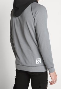 Sudadera gris con capucha negra, presenta detalles de costura, un bolsillo tipo canguro y una etiqueta de logo en el lado inferior. Diseño suave y casual.