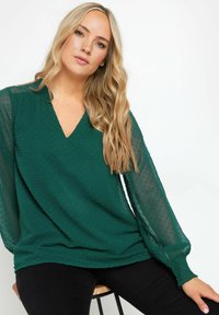 Blusa verde con scollatura a V e motivo a pois testurizzati. Maniche lunghe trasparenti con polsini arricciati, abbinata a pantaloni neri aderenti.