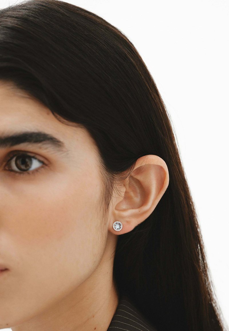 Pendiente de plata con una piedra preciosa central clara, rodeada de un borde texturizado, usado en la oreja de un modelo.