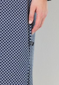 Tejido de patrón náutico con diseños geométricos blancos. Una mano sostiene el borde, revelando una pequeña etiqueta. Textura suave, forma ajustada.