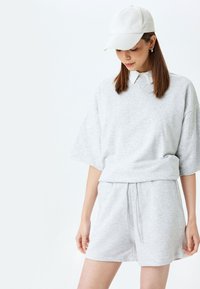 Lichtgrijze katoenen sweatshirt met korte mouwen, gecombineerd met bijpassende shorts. Het bovenstuk heeft een ronde hals en een losse pasvorm; de stof lijkt zacht. Witte baseball pet.