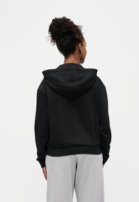 Γυναίκα με σγουρά μαλλιά δεμένα, που φοράει μαύρη κουκούλα και ανοιχτό γκρι παντελόνι, στέκεται μπροστά με την πλάτη της προς ένα απλό λευκό φόντο.