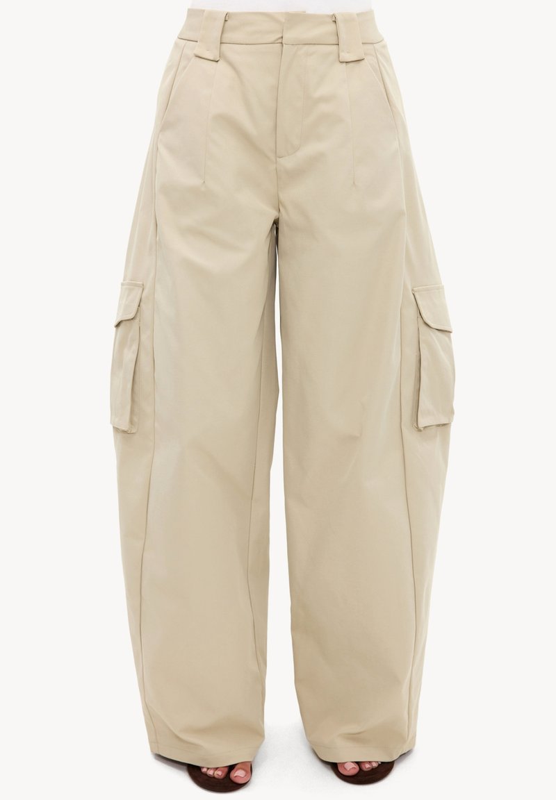 Pantalon cargo beige à jambes larges avec poches latérales à rabat, passants de ceinture et plis sur le devant, porté avec des sandales ouvertes foncées.