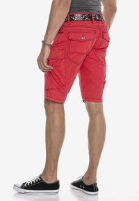 Rote Cargo-Shorts mit mehreren Taschen, sichtbaren Nähten und einem schwarzen Nieten-Gürtel. Getragen mit schwarz-weißen Turnschuhen.