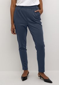 Femme portant un pantalon tapered bleu marine avec poches, associé à des escarpins noirs à talon ouvert et un haut blanc rentré, se tenant devant un fond uni.