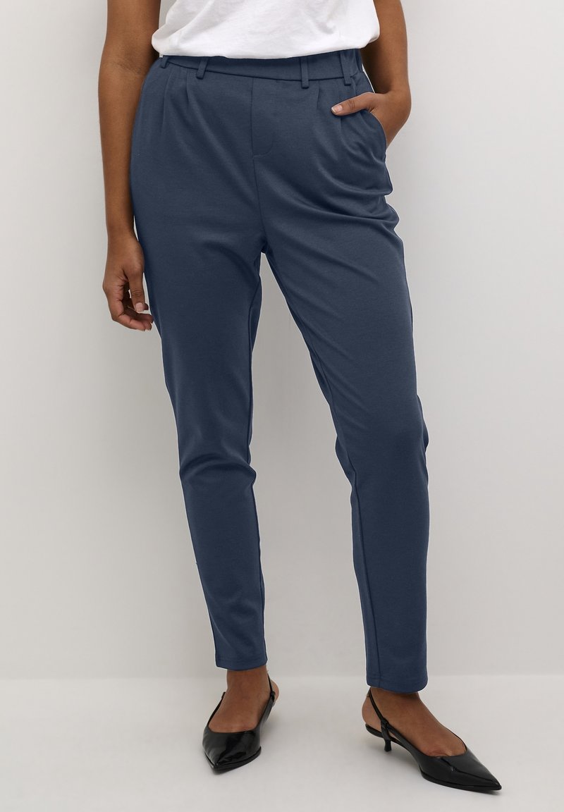 Femme portant un pantalon tapered bleu marine avec poches, associé à des escarpins noirs à talon ouvert et un haut blanc rentré, se tenant devant un fond uni.