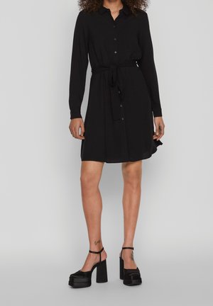 Kobieta w długiej czarnej sukience z guzikami i paskiem w talii, nosząca czarne platformowe szpilki, stojąca na tle jednolitego jasnego tła.