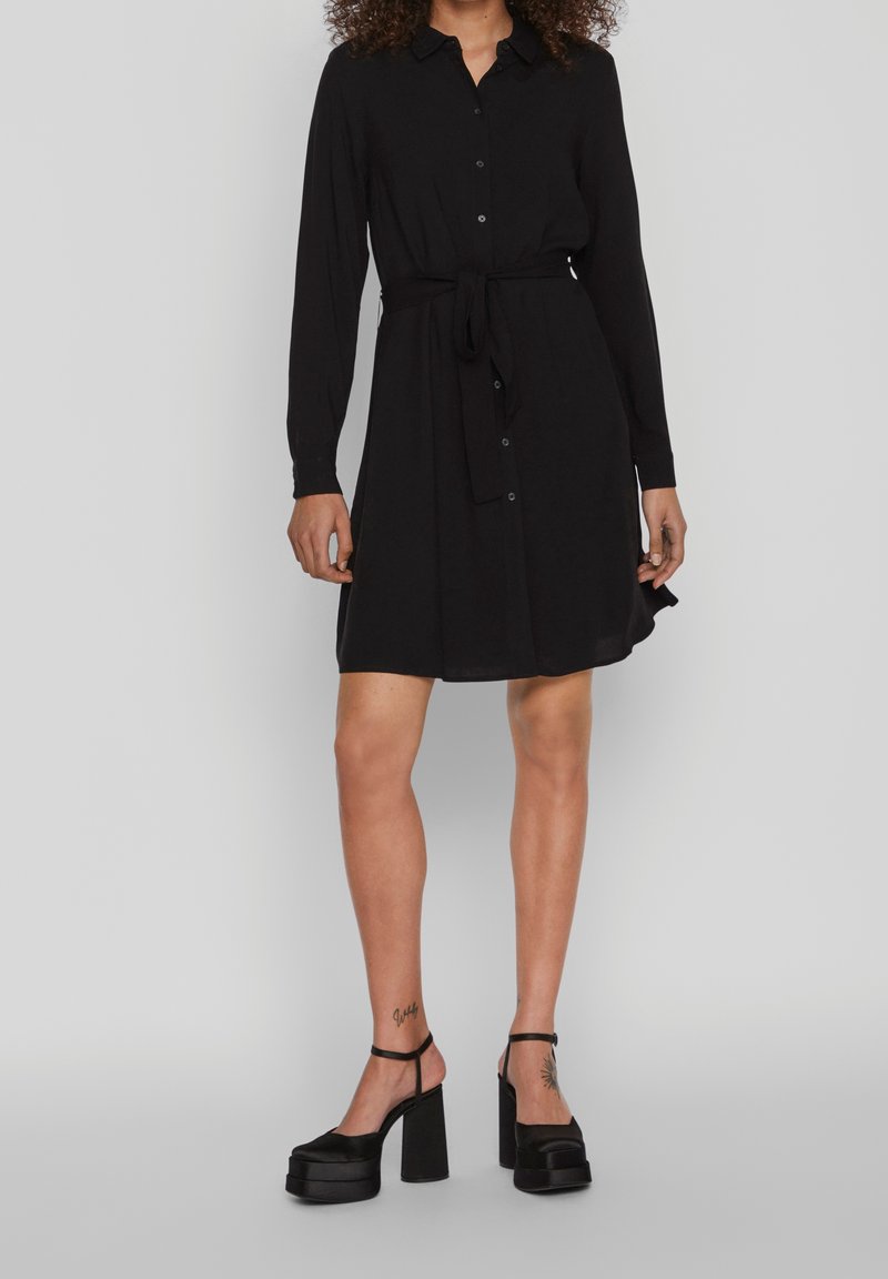 Femme en robe noire à manches longues boutonnée avec ceinture à la taille, portant des talons compensés noirs, debout devant un fond clair uni.