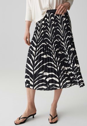 Femme portant une jupe midi plissée noire et blanche avec des motifs abstraits de feuilles, associée à des sandales à talons noires et un haut blanc.
