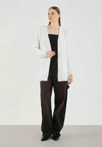 Cardigan blanco y esponjoso con frente abierto, combinado con una blusa negra y pantalones negros holgados; diseño elegante y moderno con tejido texturizado.